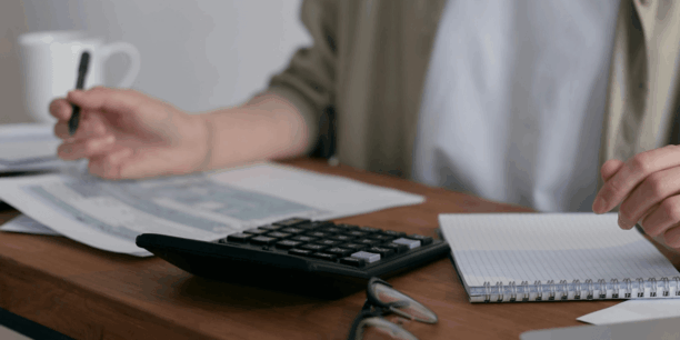 person holding a pen over a piece of paper with a calculator and notepad on the desk next to them