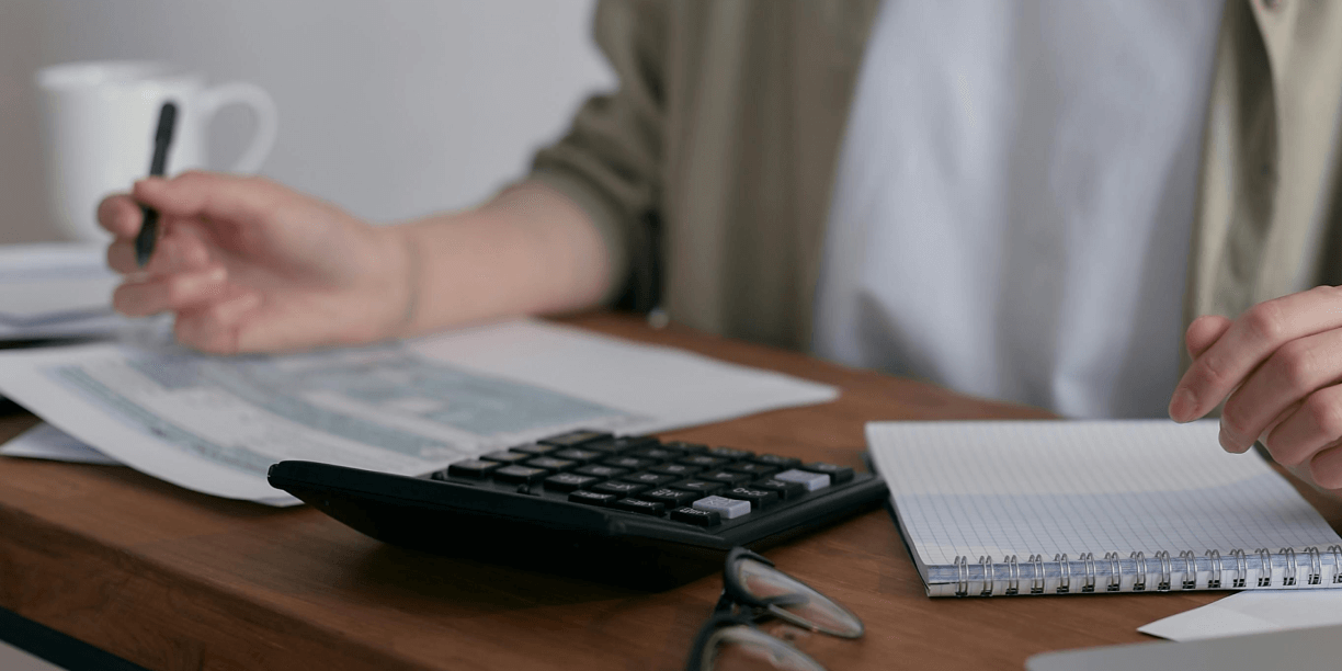 person holding a pen over a piece of paper with a calculator and notepad on the desk next to them