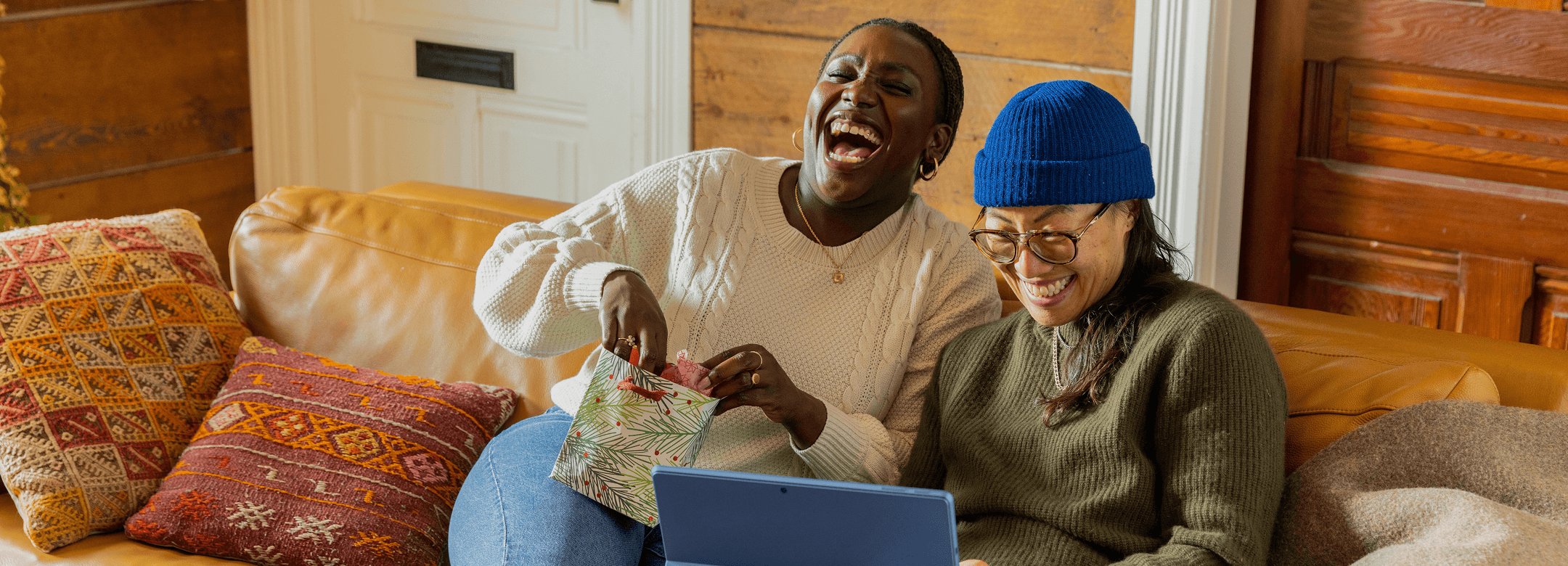 Two women laughing together on a couch while opening a gift bag and looking at a laptop in a cozy, wood-paneled living room.