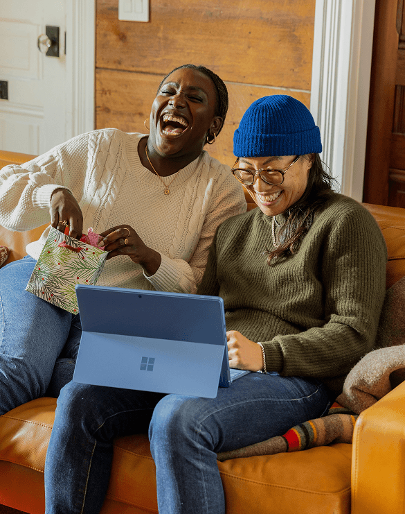 Two women laughing together on a couch while opening a gift bag and looking at a laptop in a cozy, wood-paneled living room.