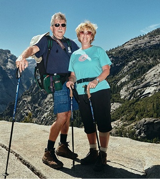 Older couple in sunglasses stand smiling on a granite overlook, each holding a trekking pole, with a rocky mountain valley behind them under a clear blue sky.