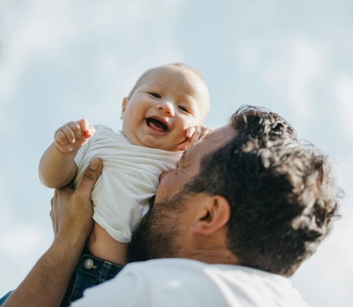 Laughing baby in a white onesie is held up in the air by a bearded man in a white shirt, against a soft blue sky with clouds.