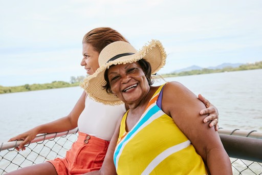 Two women lean together at the railing of a boat on calm water. The woman in front smiles broadly in a straw hat and a yellow, blue and white striped top.